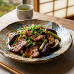 A appetizing plate of Japanese-style miso stir-fried sliced pork belly, soft eggplant, and scallions coated in a caramelized miso sauce, served on a ceramic plate alongside a bowl of steamed white rice on a rustic wooden table.