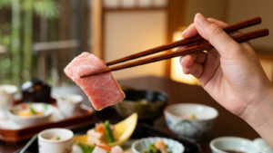 A close-up photograph of a hand gracefully holding wooden chopsticks, picking up a piece of fresh, beautifully marbled medium-fatty bluefin tuna (chutoro) sashimi. The tuna slice displays rich pink tones and delicate fat lines. In the softly blurred background, an elegant Japanese Kaiseki multi-course meal is arranged on dark wooden tables, including small ceramic dishes, soup bowls, and a peaceful view of a traditional bamboo garden through shoji screen windows.